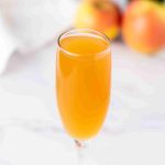 A flute glass filled with orange-colored Apple Cider Mimosa sits on a white surface, with blurred apples and a green bottle in the background.