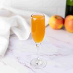 A champagne flute filled with orange-colored Apple Cider Mimosa sits on a marble surface, with two apples and a bottle in the blurred background. A white cloth is draped nearby.