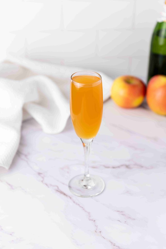 A champagne flute filled with orange-colored Apple Cider Mimosa sits on a marble surface, with two apples and a bottle in the blurred background. A white cloth is draped nearby.