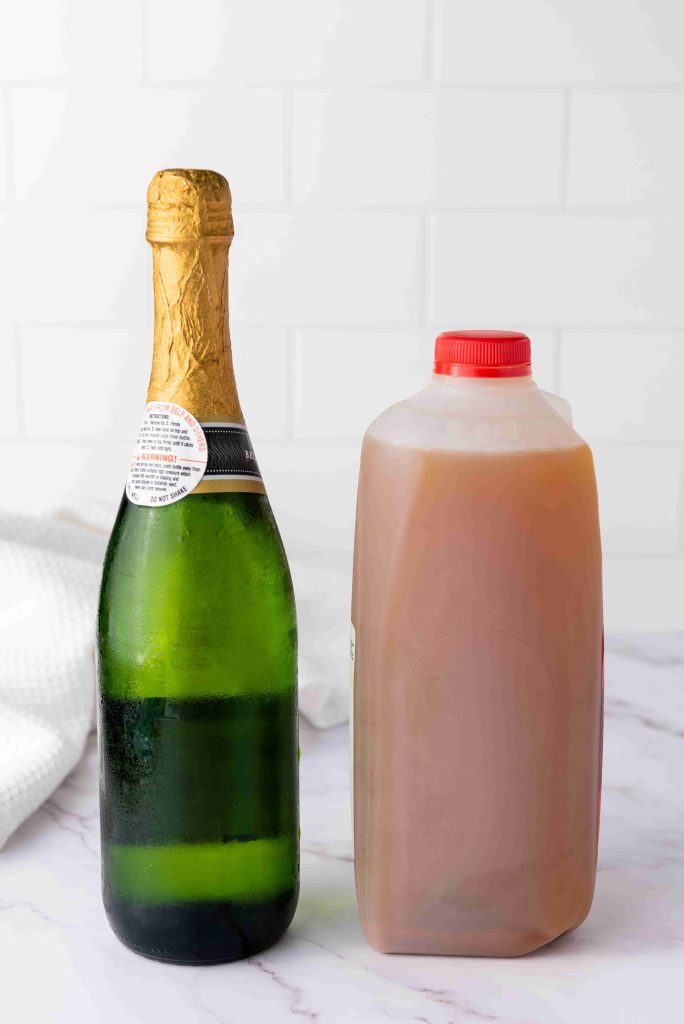 A bottle of champagne with a gold foil top stands next to a plastic jug of apple cider with a red cap, perfect for making an Apple Cider Mimosa, set on a white marble surface with a white tile background.