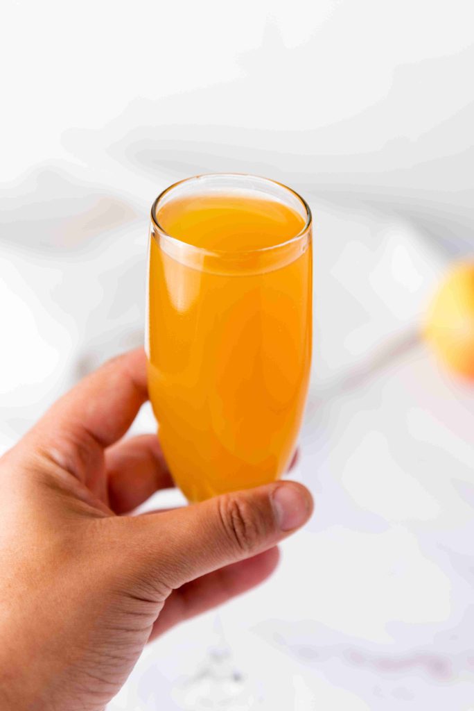 A hand holding a glass of orange-colored Apple Cider Mimosa against a blurred white background.