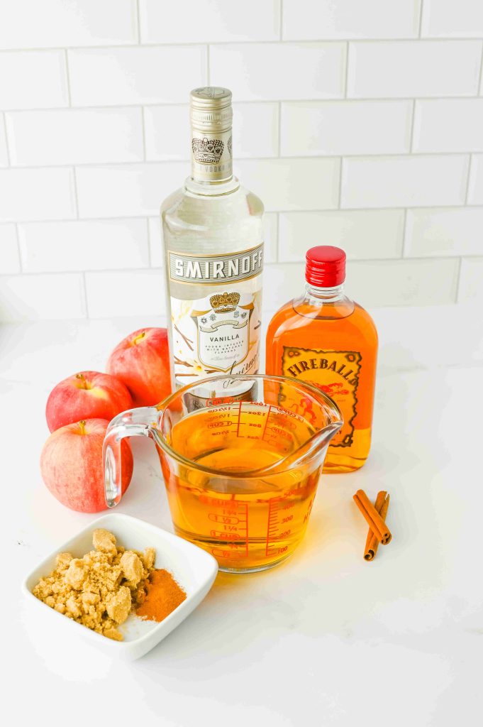 A bottle of Smirnoff vanilla vodka, Fireball whisky, apples, a measuring cup of apple cider, ground spices, and cinnamon sticks on a white surface—perfect ingredients for an apple pie cocktail fireball.