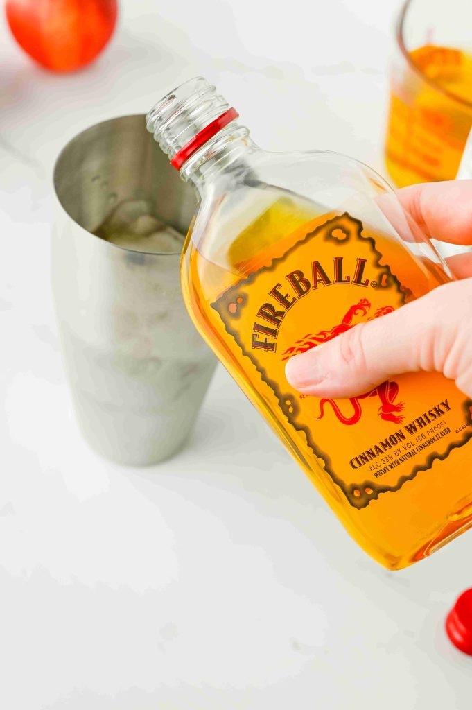 A hand pours Fireball Cinnamon Whisky from a bottle into a metal cocktail shaker filled with ice, ready to craft a delicious apple pie cocktail. In the background, a glass of whisky and an apple complete the cozy scene.