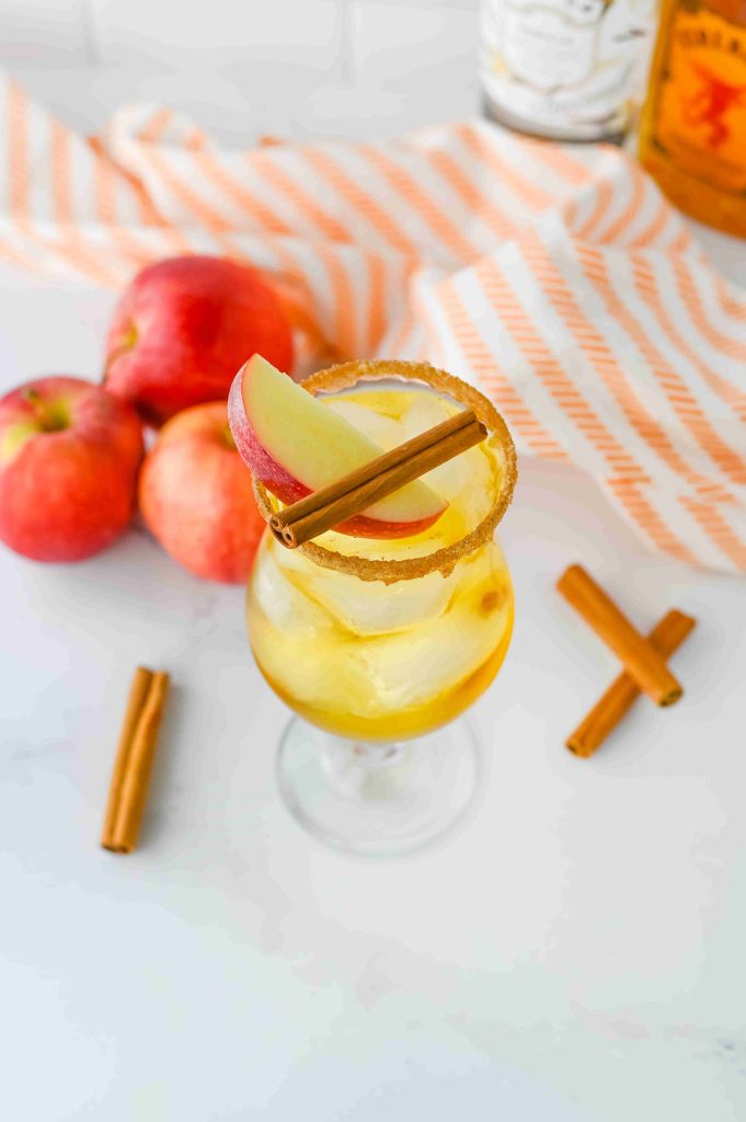 An apple pie cocktail with Fireball is garnished with apple slices and a cinnamon stick, sitting on a white surface surrounded by whole apples, cinnamon sticks, and a striped orange cloth. The glass has a sugar rim and is filled with ice.