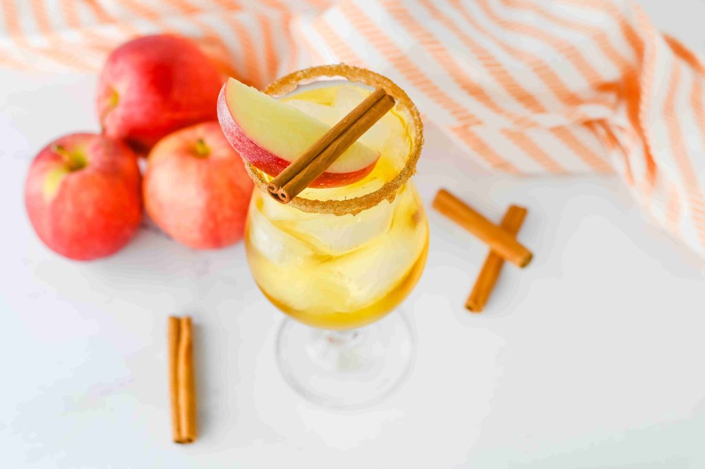 A glass of iced apple pie cocktail garnished with apple slices and a cinnamon stick, rimmed with sugar. Three apples, cinnamon sticks, and an orange-striped cloth are in the background on a white surface. Perfect for Fireball fans!.