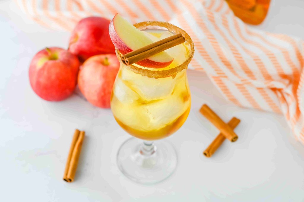 A glass filled with an iced apple pie cocktail, garnished with apple slices and a cinnamon stick. Three red apples, loose cinnamon sticks, and an orange-and-white striped cloth are in the background—perfect for pairing with Fireball.