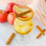 A glass of iced apple pie cocktail garnished with apple slices and a cinnamon stick, with a brown sugar rim. Fresh apples, cinnamon sticks, and a striped cloth are in the background on a white surface.