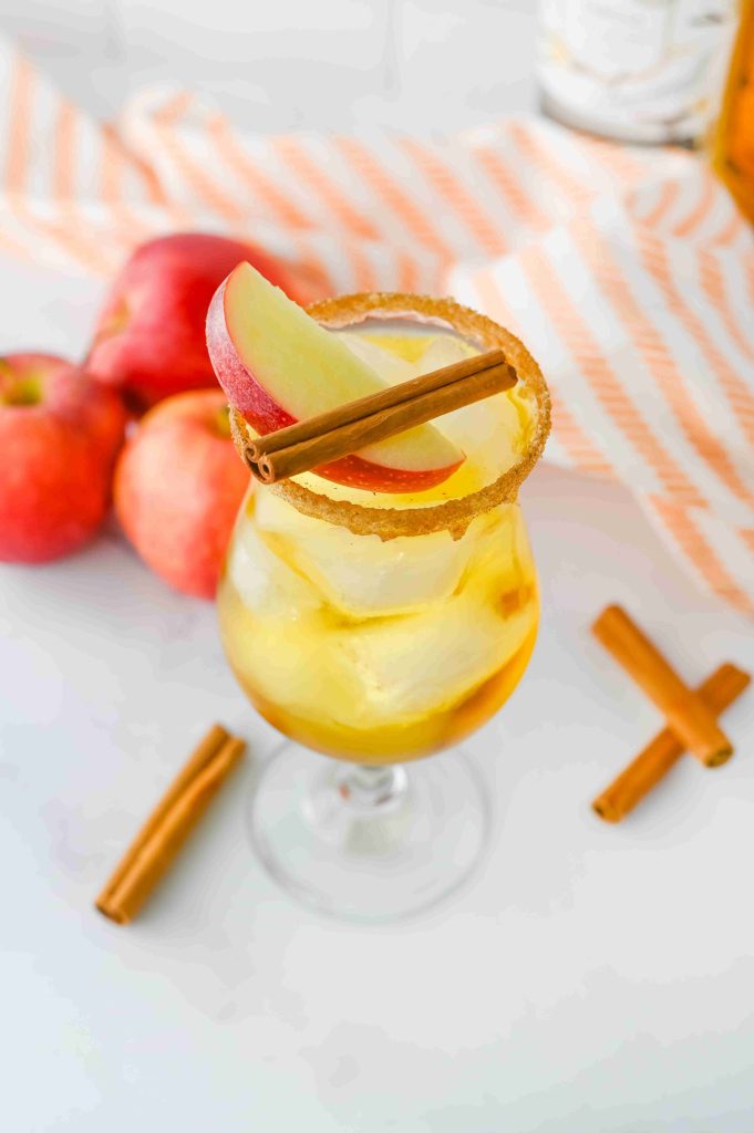 A glass of iced apple pie cocktail garnished with apple slices and a cinnamon stick, with a brown sugar rim. Fresh apples, cinnamon sticks, and a striped cloth are in the background on a white surface.