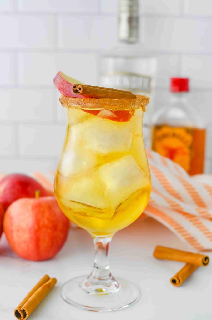 A glass of iced apple pie cocktail garnished with an apple slice and cinnamon stick, with two apples, cinnamon sticks, and bottles of Fireball in the background on a white surface.