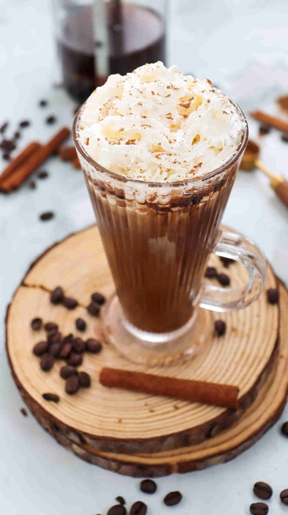 A glass mug of iced cinnamon almond milk latte topped with whipped cream and sprinkled with cinnamon sits on a wooden coaster, surrounded by coffee beans and cinnamon sticks on a white surface.