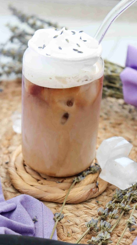 A glass of lavender iced coffee topped with whipped cream and lavender buds, served with a clear straw. The drink sits on a woven coaster, surrounded by lavender sprigs and purple cloth.