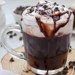 A glass mug of dairy-free hot chocolate topped with whipped cream, chocolate drizzle, and chocolate chips sits on a wooden coaster, with extra chocolate chips scattered around and a dessert in the background.