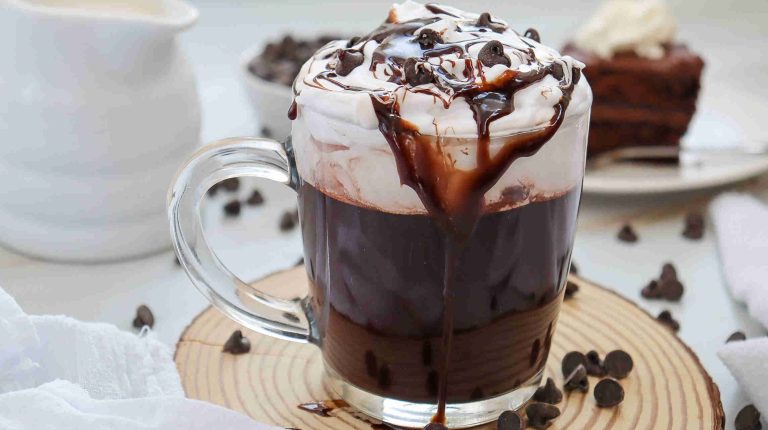 A glass mug of dairy-free hot chocolate topped with whipped cream, chocolate drizzle, and chocolate chips sits on a wooden coaster, with extra chocolate chips scattered around and a dessert in the background.