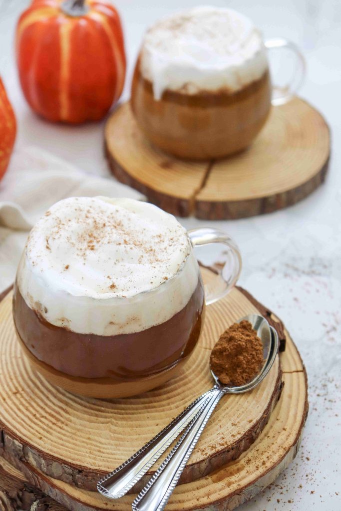 Two glass mugs of frothy dairy free pumpkin spice latte sit on wooden coasters, with a spoonful of ground spices beside them. An orange pumpkin decorates the background on a light, textured surface.