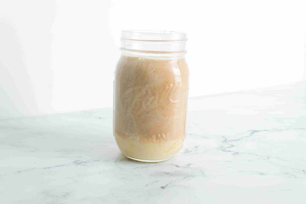 A glass mason jar filled with a light brown smoothie or blended drink, featuring pumpkin spice coffee creamer, sits on a white marble surface against a plain white background.