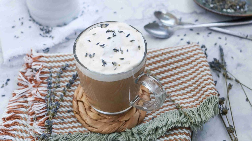 A glass mug of creamy earl grey milk tea topped with foam and lavender buds sits on a woven coaster atop a striped, fringed cloth, with lavender sprigs and two spoons nearby.