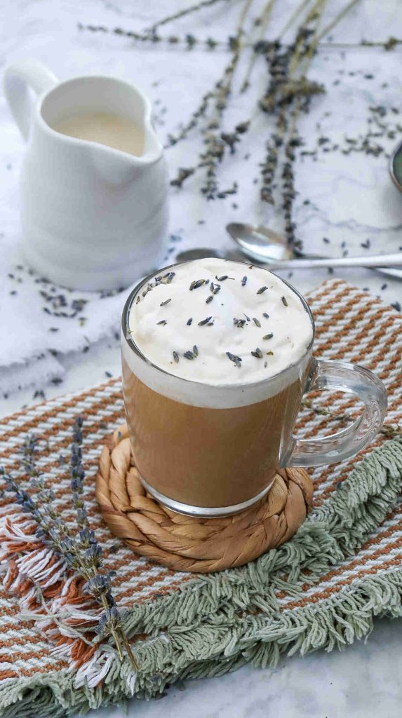 A glass mug of earl grey milk tea topped with creamy foam and lavender buds sits on a woven coaster, with a small white pitcher and sprigs of dried lavender nearby on a textured cloth and marble surface.