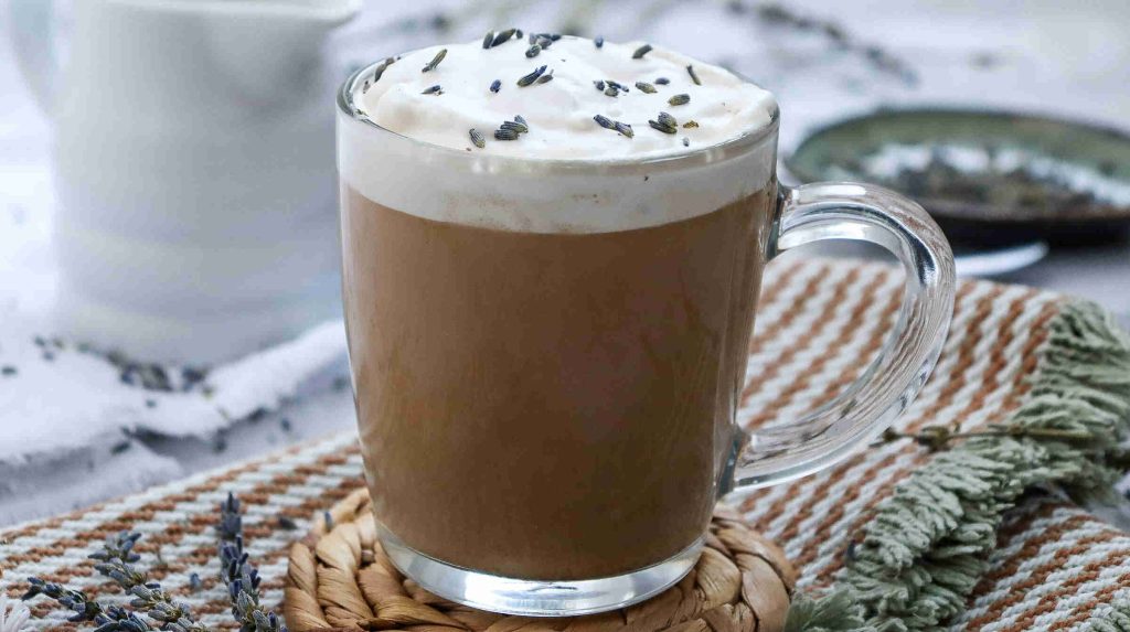 A glass mug filled with creamy earl grey milk tea topped with whipped cream and sprinkled lavender buds, set on a woven coaster with a striped cloth and lavender sprigs nearby.