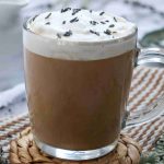 A glass mug filled with creamy earl grey milk tea topped with whipped cream and sprinkled lavender buds, set on a woven coaster with a striped cloth and lavender sprigs nearby.