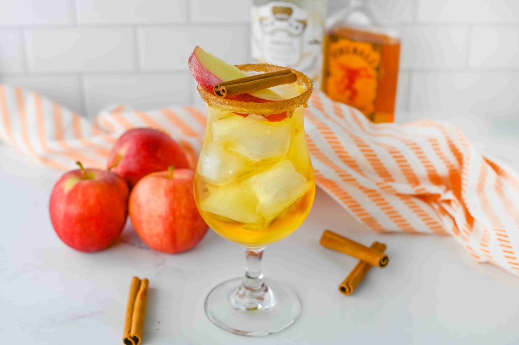 A glass of iced apple pie cocktail with a cinnamon stick and apple slice garnish, surrounded by red apples and cinnamon sticks, on a white surface with orange-striped cloth and Fireball bottles in the background.