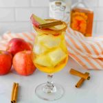 A glass of iced apple pie cocktail with a cinnamon stick and apple slice garnish, surrounded by red apples and cinnamon sticks, on a white surface with orange-striped cloth and Fireball bottles in the background.