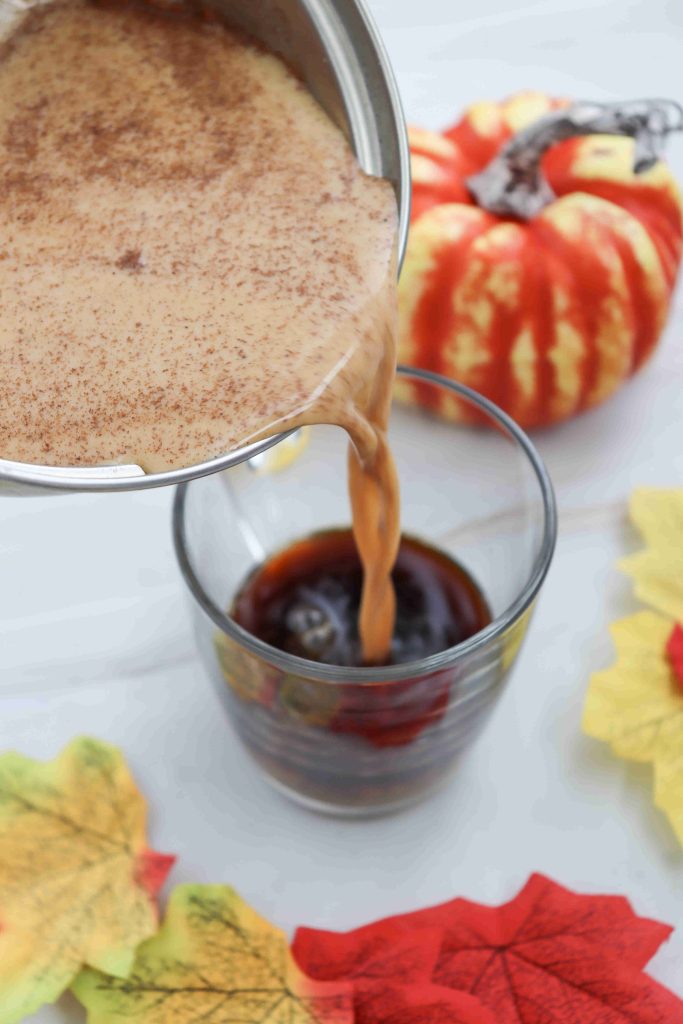 A creamy, cinnamon-spiced mixture for a pumpkin spice latte with almond milk is being poured from a pot into a glass of iced coffee. Autumn leaves and a decorative pumpkin are arranged nearby on a white surface.