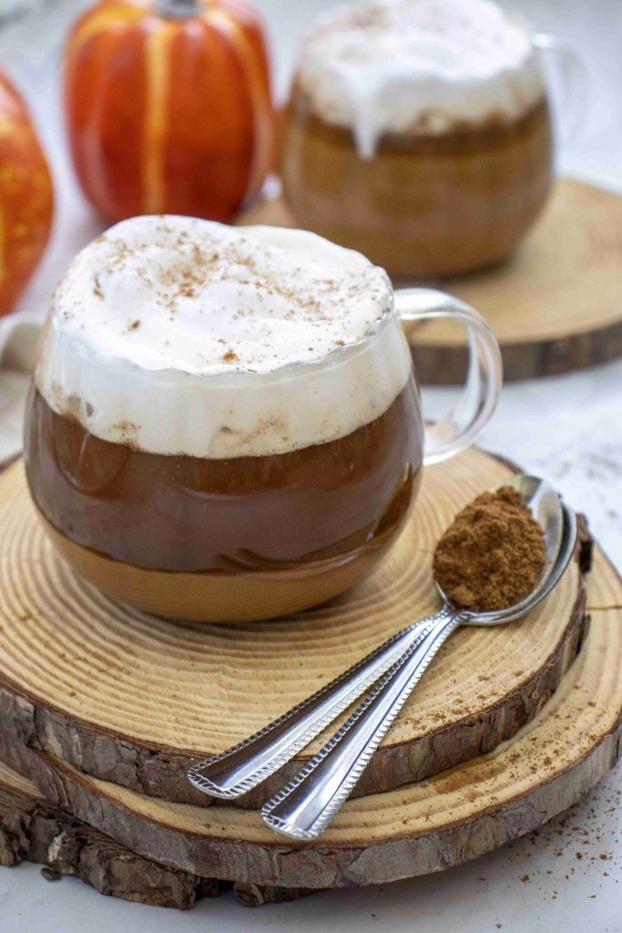 A glass mug of dairy free pumpkin spice latte topped with whipped cream and cinnamon sits on a wooden coaster, with two spoons filled with spices nearby. A second mug and small pumpkins are in the background.