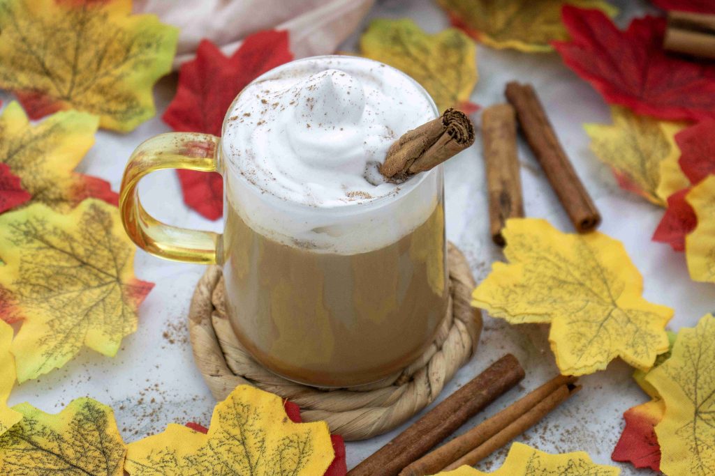 A pumpkin spice latte with almond milk topped with whipped cream and a cinnamon stick sits on a coaster, surrounded by cinnamon sticks and colorful autumn leaves.