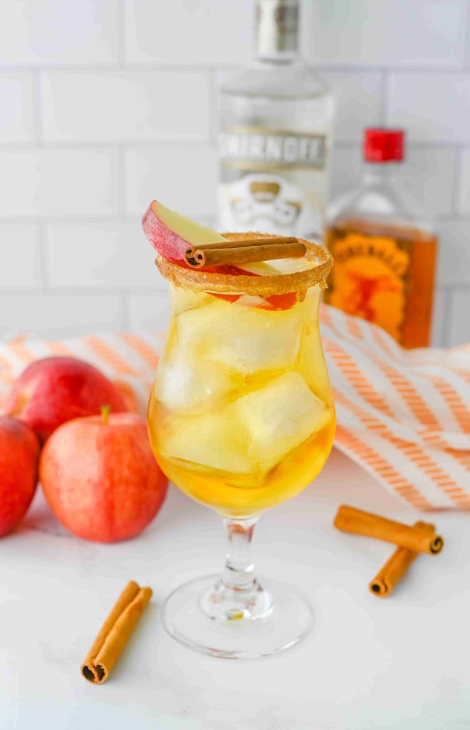 A cocktail glass filled with ice and a golden apple pie cocktail, garnished with a red apple slice and cinnamon stick. Bottles of Fireball, apples, and cinnamon sticks are in the background on a white surface.