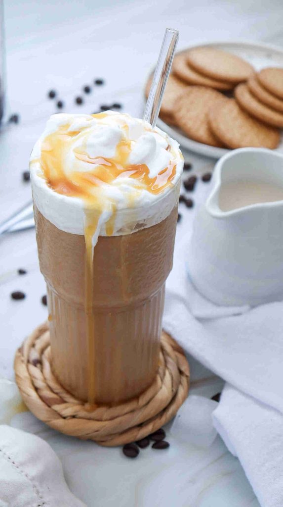 A glass of iced coffee topped with whipped cream and caramel drizzle, some caramel dripping down the side—just like in guides on how to make caramel iced coffee at home. In the background are cookies, a white jug, and scattered coffee beans.