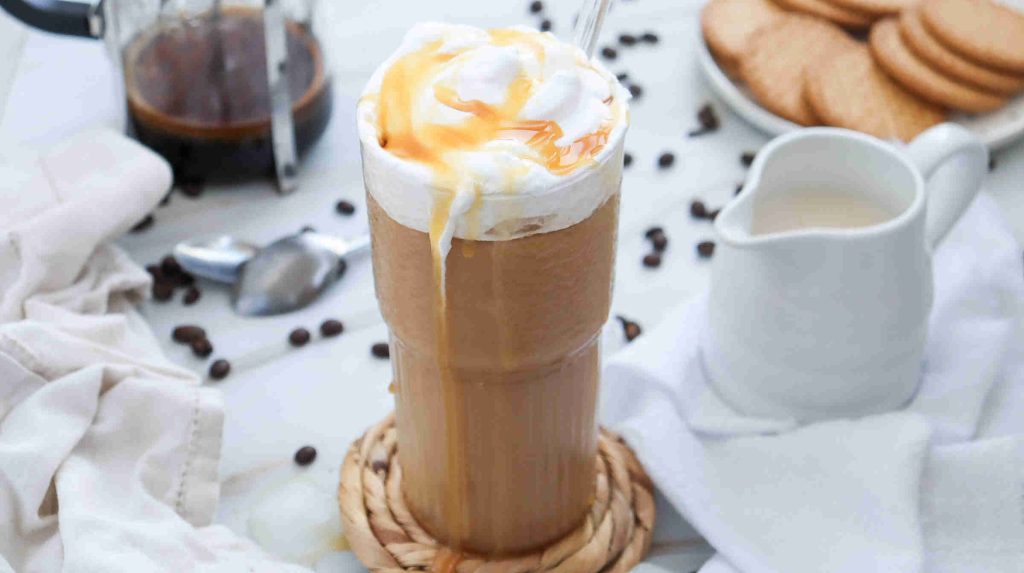 A tall glass of iced coffee topped with whipped cream and caramel drizzle sits on a woven coaster, inviting you to discover how to make caramel iced coffee at home. Coffee beans, cookies, and a white creamer pitcher complete the cozy scene.