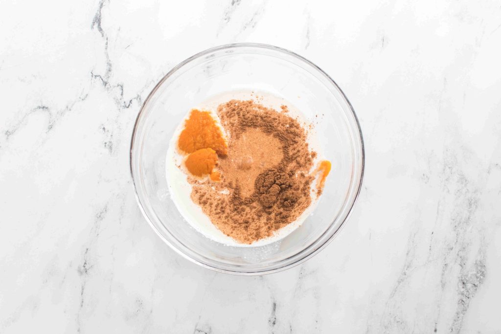 A glass bowl on a white marble surface contains milk, pumpkin puree, and brown spices, ready to be mixed into a homemade pumpkin spice coffee creamer.