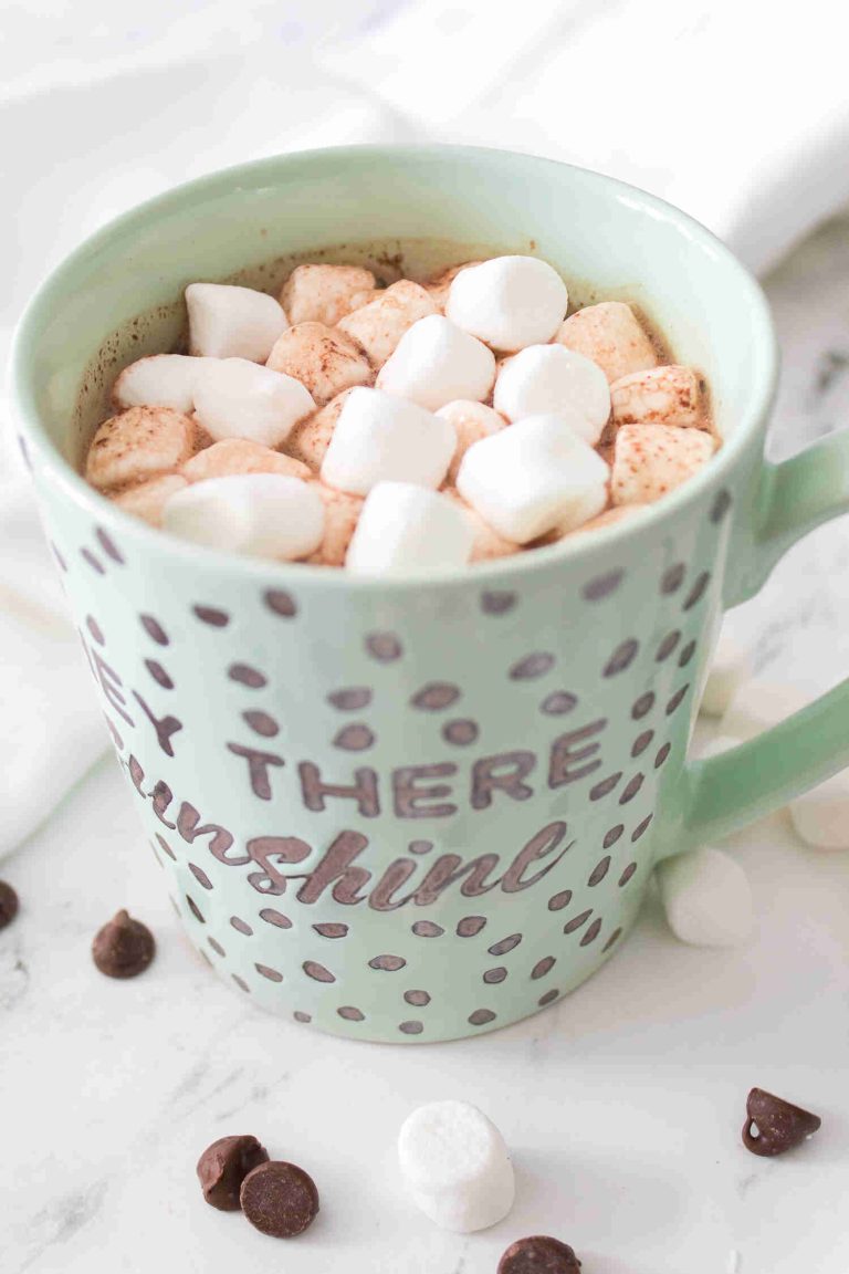 A mint green mug filled with homemade hot chocolate with chocolate chips, topped with mini marshmallows, sits on a white surface scattered with chocolate chips and marshmallows. The polka dot mug features the words Hey There Sunshine.