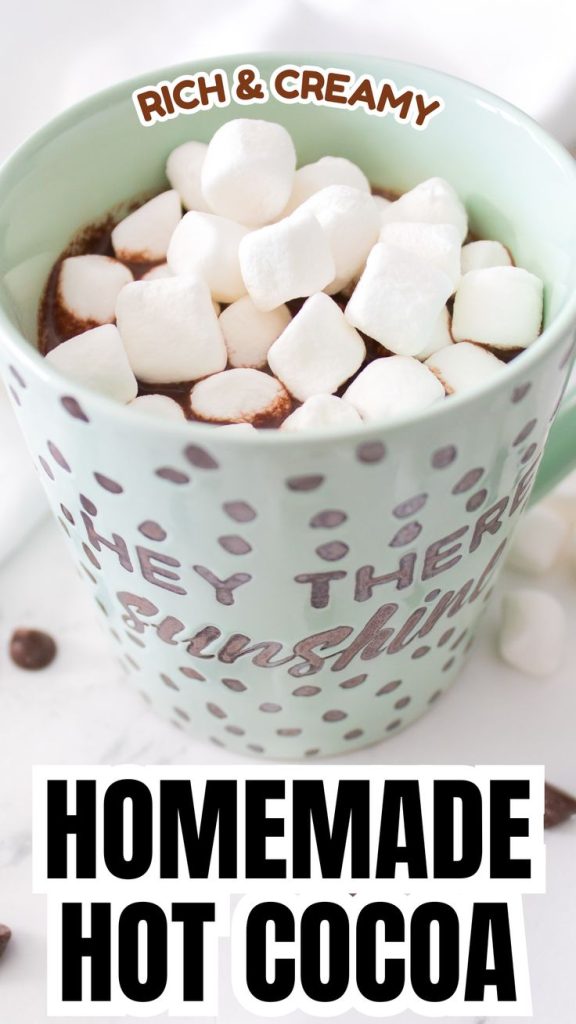 A light green mug filled with homemade hot chocolate with chocolate chips, topped with mini marshmallows. The mug has a spotted design and the phrase HEY THERE sunshine. Text on the image reads RICH & CREAMY and HOMEMADE HOT COCOA.