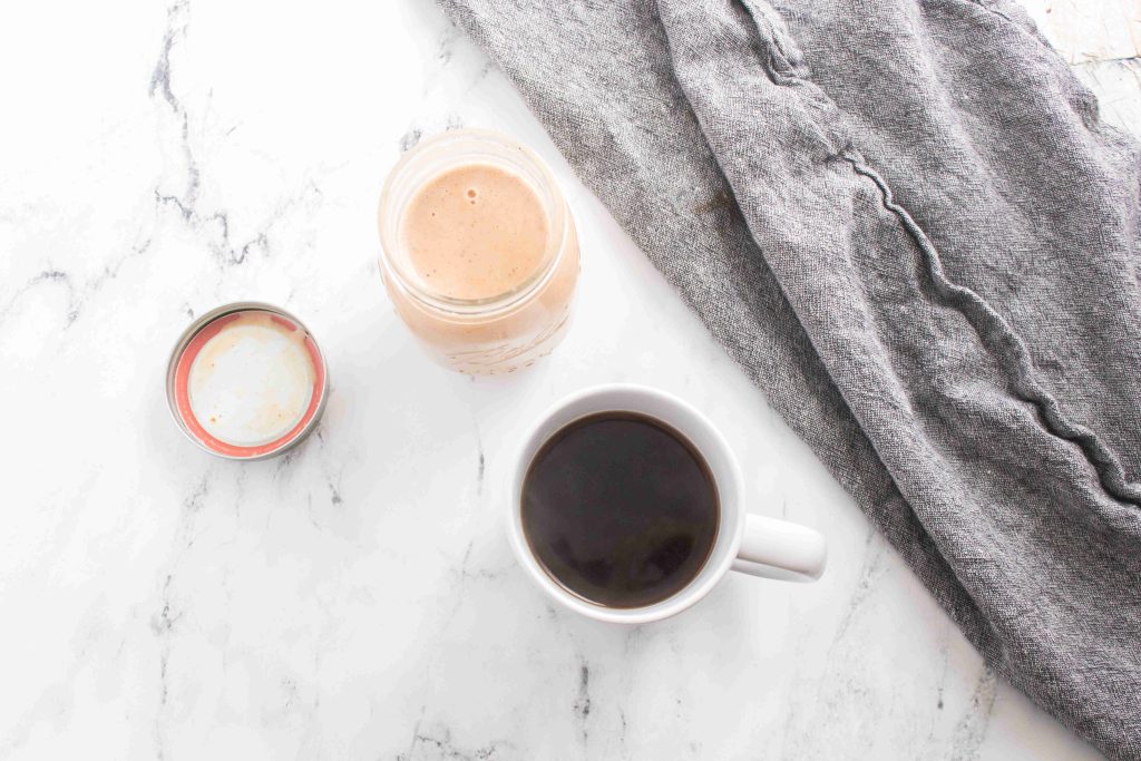 A cup of black coffee, an open jar with creamy brown pumpkin spice coffee creamer, and a gray cloth napkin rest on a white marble surface.