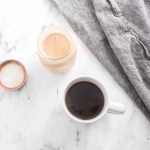 A cup of black coffee, an open jar with creamy brown pumpkin spice coffee creamer, and a gray cloth napkin rest on a white marble surface.