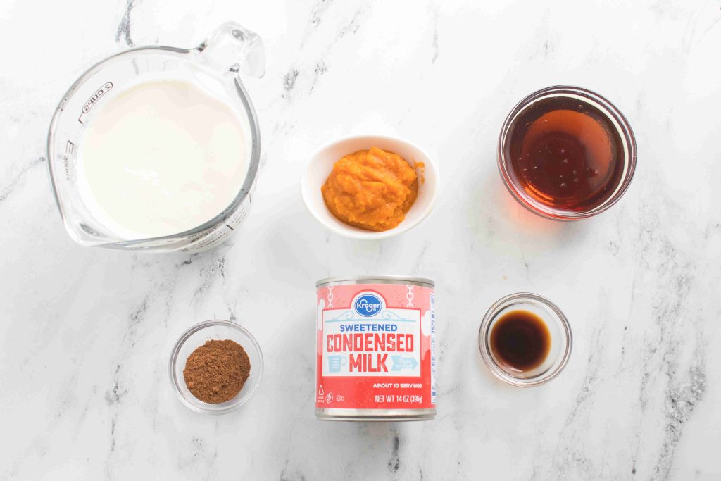 Six ingredients for pumpkin spice coffee creamer on a marble surface: heavy cream, pumpkin puree, maple syrup, cocoa powder, sweetened condensed milk, and vanilla extract, each displayed in separate bowls or containers.