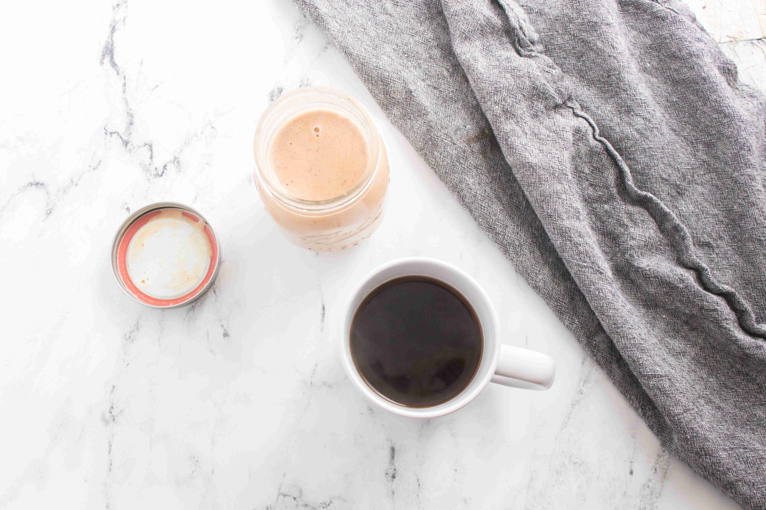 A cup of black coffee, an open jar with creamy brown pumpkin spice coffee creamer, and a gray cloth napkin rest on a white marble surface.