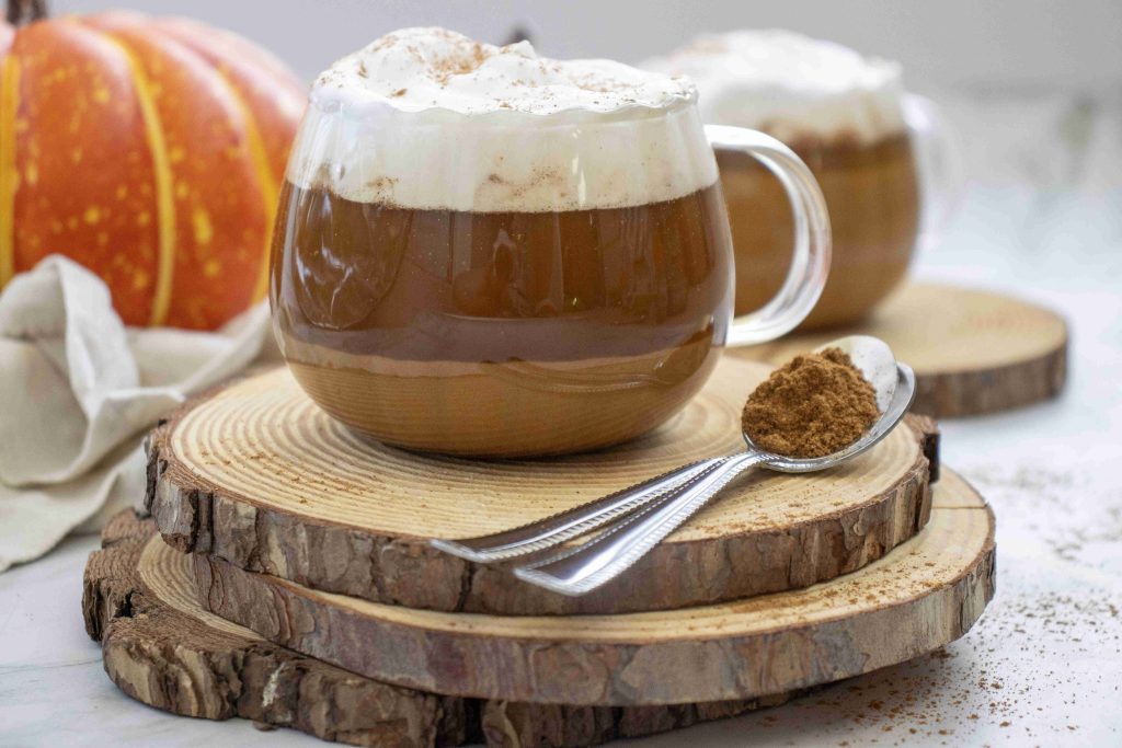 A glass mug filled with dairy free pumpkin spice latte, topped with whipped cream and sprinkled cinnamon, sits on a wooden coaster slice. A spoon with ground cinnamon and a pumpkin are in the background.