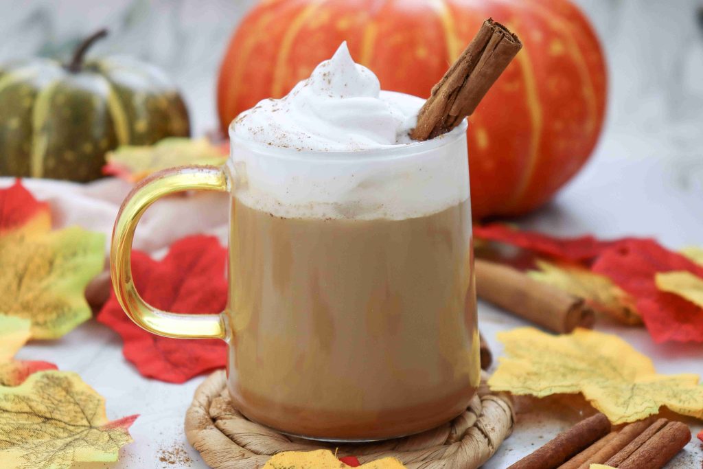 A glass mug of pumpkin spice latte with almond milk, topped with whipped cream and a cinnamon stick, sits on a coaster, surrounded by autumn leaves, cinnamon sticks, and pumpkins in the background.