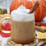 A glass mug of pumpkin spice latte with almond milk, topped with whipped cream and a cinnamon stick, sits on a coaster, surrounded by autumn leaves, cinnamon sticks, and pumpkins in the background.