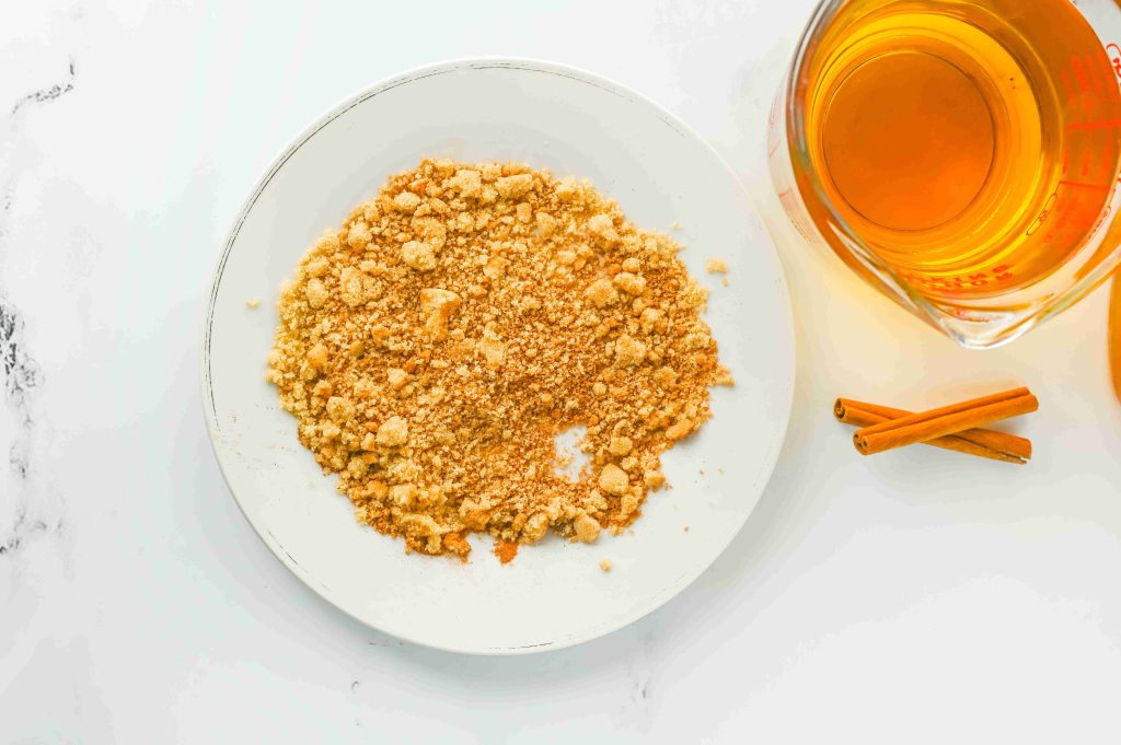 A white plate with crumbled graham crackers sits next to a glass measuring cup filled with golden liquid and two cinnamon sticks—perfect for crafting an apple pie cocktail with a splash of Fireball on a white surface.