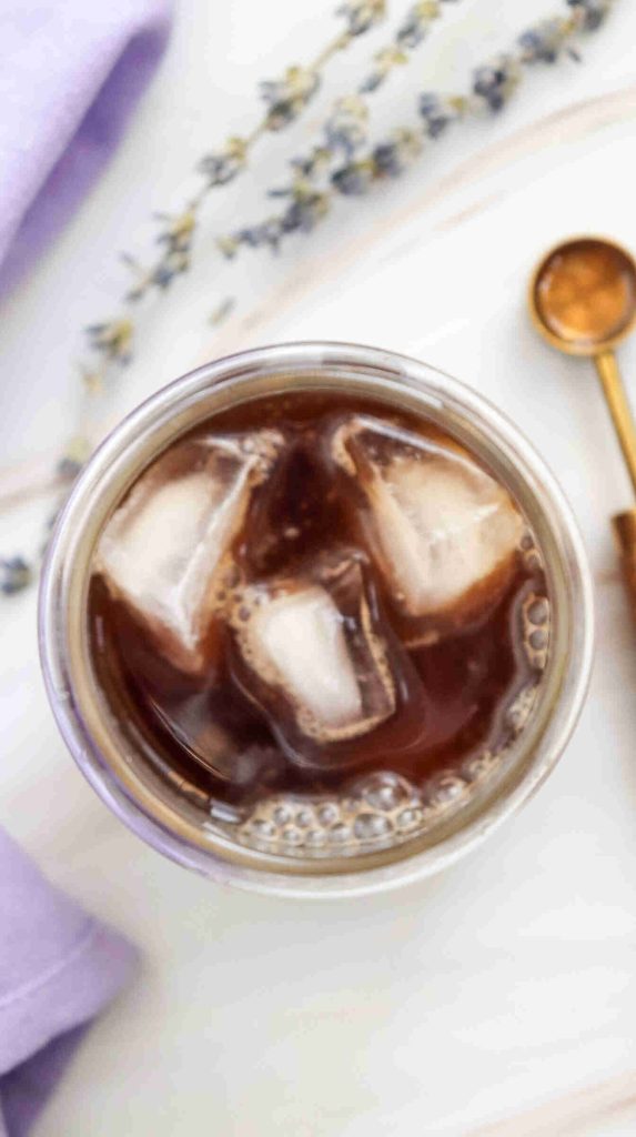 A top-down view of a glass of lavender iced coffee with large ice cubes, next to a gold spoon, a lavender sprig, and a folded lavender napkin on a white surface.