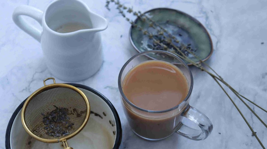 A glass mug of earl grey milk tea sits on a marble surface next to a white pitcher of milk, a strainer with tea leaves, a green dish, and a sprig of dried herbs.