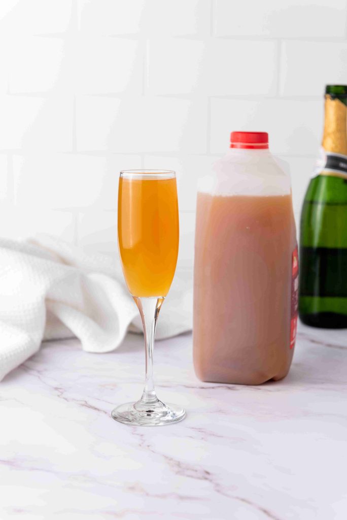A flute glass filled with a light orange Apple Cider Mimosa sits on a marble surface next to a plastic jug of apple cider and a bottle of champagne, with a white cloth in the background.
