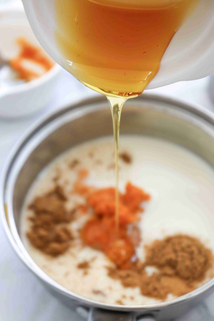 Honey being poured from a white bowl into a pot containing milk, pumpkin puree, and spices—perfect for preparing a cozy dairy free pumpkin spice latte. A blurred bowl with ingredients sits in the background.