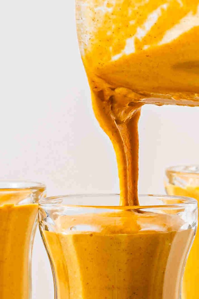 A thick, creamy pumpkin pie spice milkshake is being poured from a blender into a clear glass, with two other filled glasses nearby.