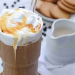 A tall glass of iced coffee topped with whipped cream and caramel sauce, with a straw—perfect inspiration for how to make caramel iced coffee at home. In the background, cookies, a jug of milk, coffee beans, and a spoon rest on a white surface.