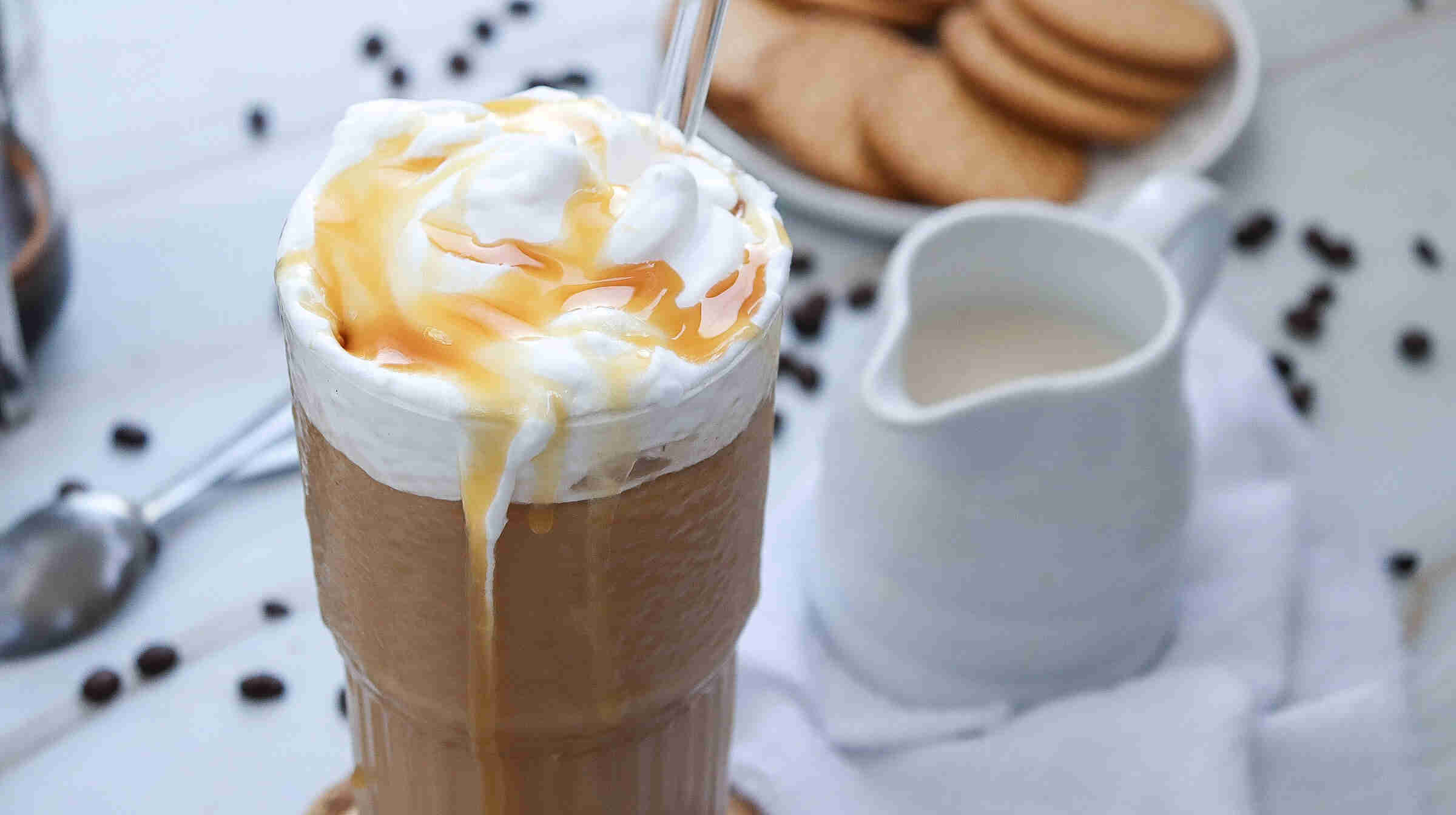 A tall glass of iced coffee topped with whipped cream and caramel sauce, with a straw—perfect inspiration for how to make caramel iced coffee at home. In the background, cookies, a jug of milk, coffee beans, and a spoon rest on a white surface.
