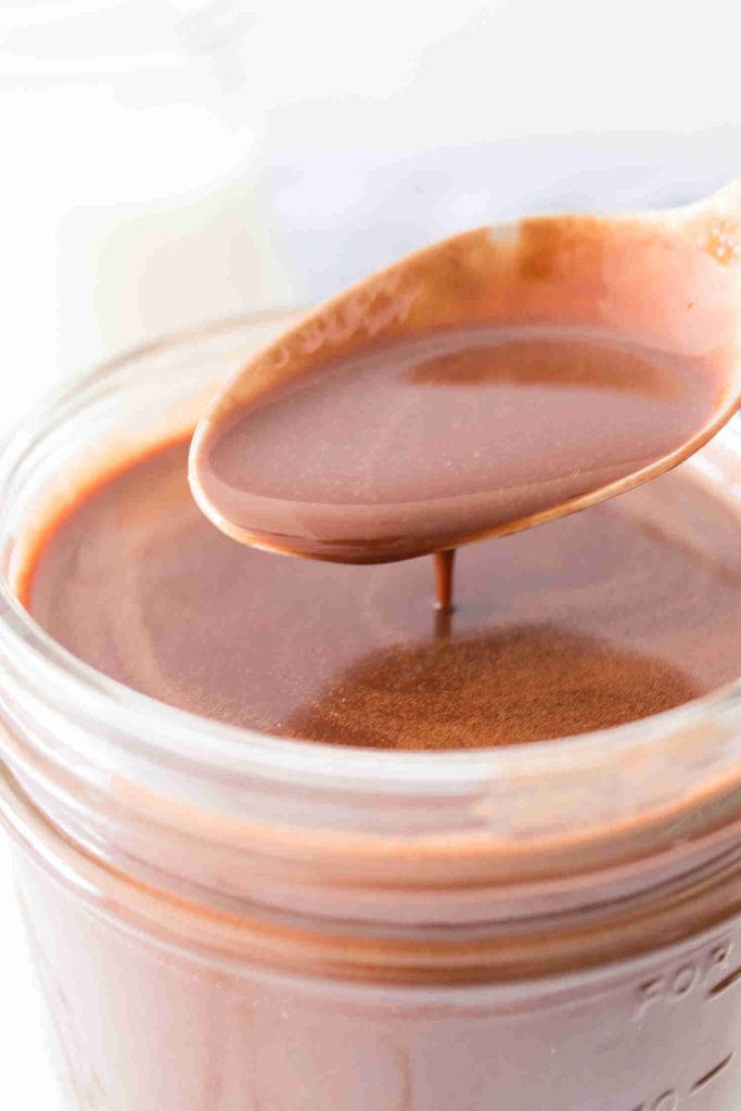 A close-up of a spoon dripping smooth, melted chocolate over a glass jar filled with rich cocoa syrup.